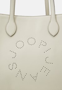 Sac fourre-tout en cuir beige avec un design de lettrage perforé. Deux longues poignées et surface texturée. Le corps principal est lisse avec un grain subtil.