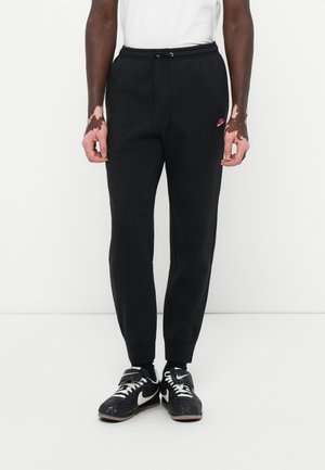 Mężczyzna ubrany w czarne spodnie dresowe Nike i czarne buty Nike z białymi logo Swoosh, stojący na jednolitym białym tle.