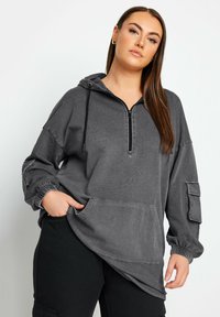 Grå huvtröja med dragkedja i nacken, ribbad struktur, stor framficka och elastiska muddar. Har sidofickor på ärmarna.