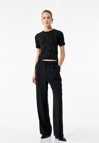 Sort ribbet strikbluse med korte ærmer, udsmykket med perle-lignende detaljer. Sammen med højtaljede sorte bukser med brede ben og elegante sorte hæle.