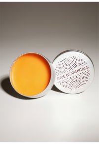 Eine runde silberne Dose mit einem leuchtend orangefarbenen Balsam. Der Deckel hat ein gepunktetes Muster und den Text "TRUE BOTANICALS" in Burgundy.