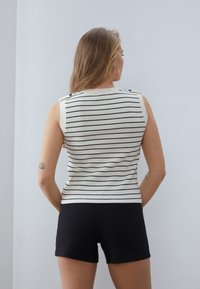 Gestreiftes ärmelloses Oberteil in Weiß und Schwarz mit gerippter Textur und Knopfakzenten an den Schultern, kombiniert mit schwarzen Shorts.