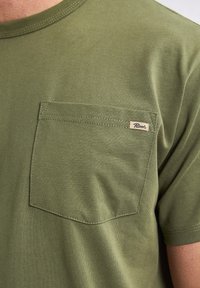 Olive green t-shirt made of soft cotton, featuring a chest pocket and a stitched logo tag. Short sleeves and a classic crew neck design.