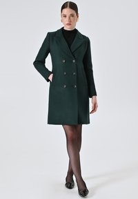 Cappotto doppiopetto verde in misto lana con due tasche frontali e colletto a poncho, caratterizzato da bottoni neri e una vestibilità strutturata e su misura.