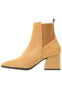 Stivaletto in suede beige con punta affusolata, pannelli laterali elasticizzati e tacco squadrato chunky. Texture liscia e dettagli di design minimi.