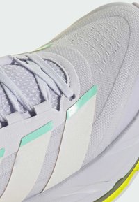 Gros plan d'une chaussure de sport blanche avec des accents vert clair, un modèle à lacets, une tige en tissu texturé et un détail jaune sur le bord de la semelle.