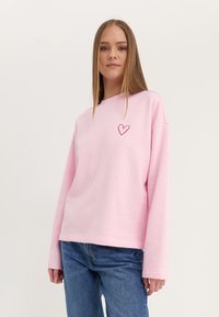 Roosa ülekaaluline sweatshirte, mis on valmistatud pehmest materjalist ja millel on pikad varrukad. Ees on väike, tikandiga südame detail.
