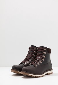 Botas de senderismo de cuero marrón con ojales de metal, cordones marrones y un acolchado en el tobillo. La suela tiene un diseño texturizado con un patrón de tracción prominente.