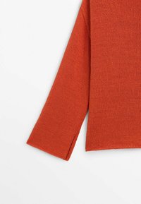 Pull en maille à manches longues de couleur orange brûlé avec un ourlet droit et une discrète fente latérale au niveau du poignet.