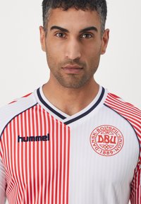 Maillot de sport rayé rouge et blanc avec un col en V, présentant un logo sur le côté gauche de la poitrine et une étiquette "hummel" sur le côté. Texture de tissu doux.