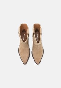 Cizme din suede bej, cu vârf ascuțit, cusături decorative și capse de prindere pe fiecare parte pentru un acces facil. Finisaj neted și talpă plată.