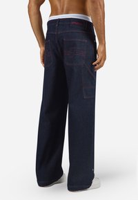 Wide-leg dark denim pants with red stitching, featuring two back pockets. Includes a white waistband and a logo at the back.