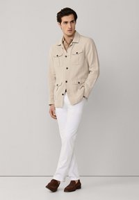 Mann trägt eine beigefarbene, geknöpfte Jacke über einem hellbraunen Hemd, weiße Hose und braune Loafers. Er steht mit einer Hand in der Tasche und überschlagenen Beinen.