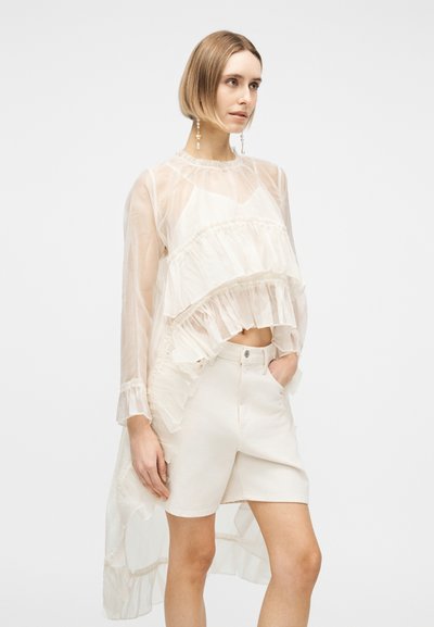 Γυναίκα που φοράει διαφανές λευκό μπλουζάκι με στρώσεις και βολάν πάνω από λευκά σορτς, με το ένα χέρι στην τσέπη και ουδέτερη έκφραση, ποζάροντας σε απλό φόντο.