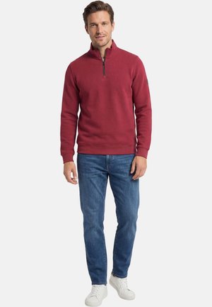 Uomo che indossa un pullover rosso con cerniera a quarto, jeans blu e sneaker bianche, in piedi davanti a uno sfondo chiaro e uniforme.
