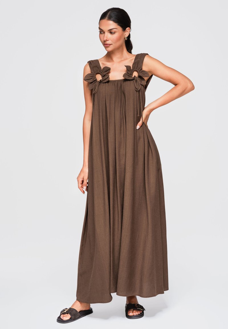 Femme portant une longue robe marron sans manches avec des liens floraux aux épaules, debout avec une main sur la hanche et des sandales noires à enfiler.