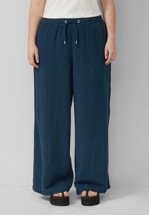 Osoba nosząca granatowe szerokie spodnie z troczkiem w pasie i czarne sandały na platformie, stojąca przy jednolitej szarej ścianie.