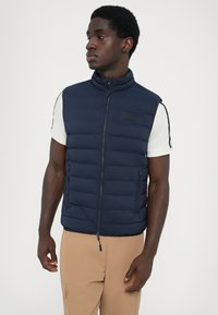 Navy puffer vest with high collar, zip closure, and side pockets. Features a textured surface and logo patch on the chest.