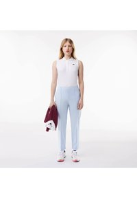 Polo senza maniche bianco con logo verde, pantaloni su misura azzurri chiari e giacca bordeaux appoggiata su un braccio. Sneakers bianche con accenti rosa.
