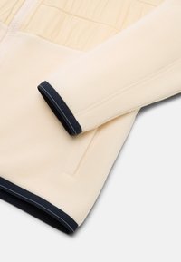 Fleecejakke i creme med ribbet marinblå manchet. Har lynlås og en rynket design langs kroppen for et tekstureret udseende.