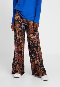 Zwarte wijde broek van stof met een veelkleurig paisley- en bloemenpatroon. Witte sneakers zichtbaar aan de onderkant.