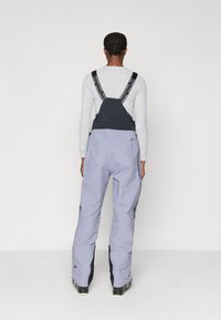 Svetlo vijolični snežni hlače z nastavljivimi črnimi naramnicami in stranskimi zadrgami. Narejene so iz trpežnega materiala, z ohlapnim krojem in dekorativnim šivanjem.