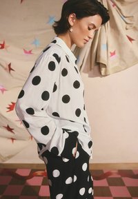 Biela košeľa s čiernymi bodkami, skombinovaná s čiernymi nohavicami s rovnakými bodkami. Ľahký materiál, voľný strih, dlhé rukávy.