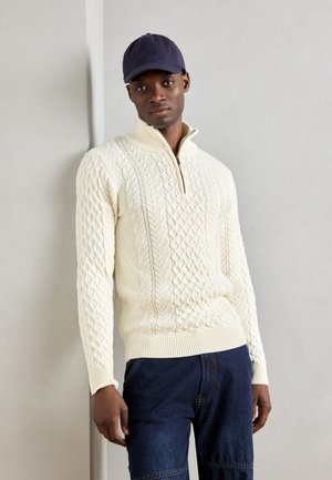 Suéter de punto trenzado color crema con cuello alto y media cremallera, combinado con pantalones de mezclilla oscuros. La tela tiene un patrón texturizado.