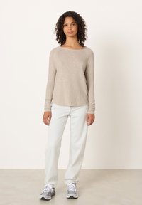 Moteris su garbanotais plaukais, dėvinti smėlio spalvos ilgomis rankovėmis marškinėlį, šviesiai mėlynas džinsus ir pilkus sportinius batelius, stovinti priešais paprastą smėlio spalvos foną.