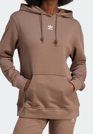 Osoba ubrana w brązową bluzę z kapturem Adidas i pasujące spodnie, ręce w kieszeni bluzy, widoczne logo Adidas na piersi i spodniach.