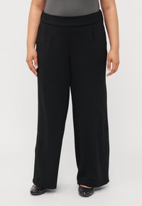 Sorte vide benbukser med flat midje og folder; glatt tekstur uten synlige mønstre; kombinert med svarte spikret slip-on sko.