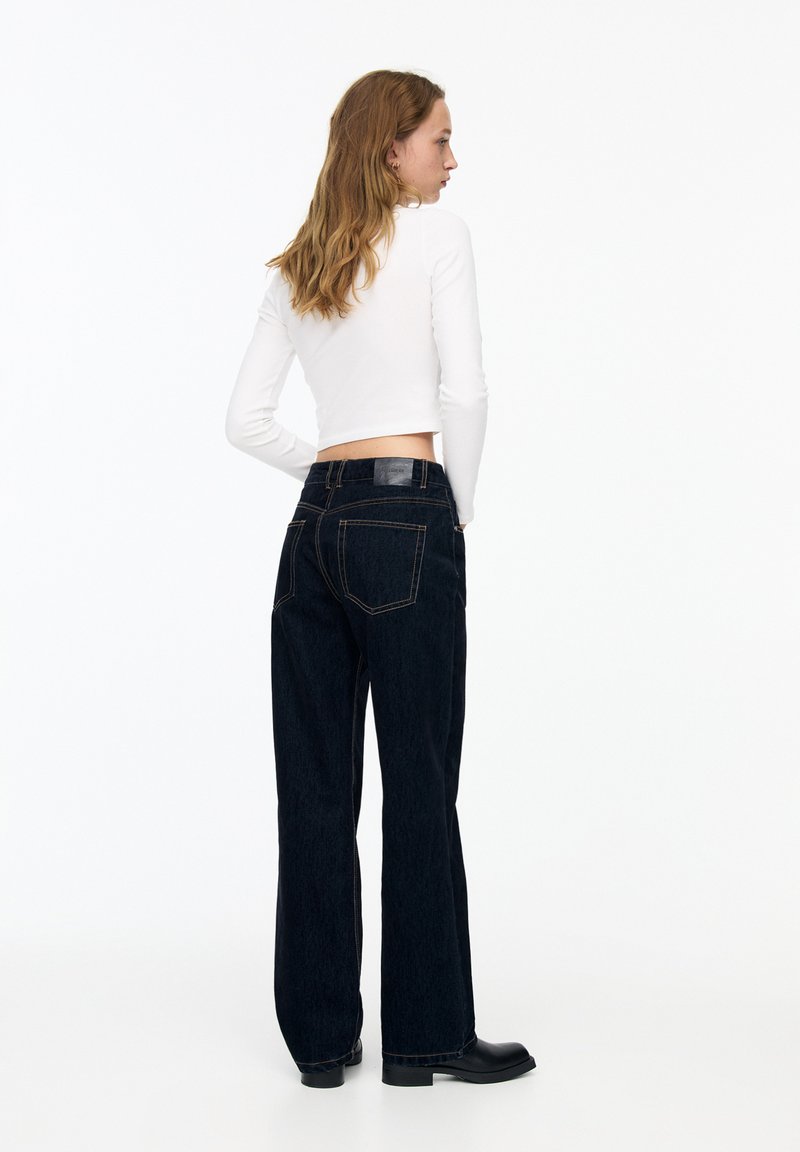 Hoch geschnittene dunkelblaue Jeans mit geradem Bein, Fünf-Taschen-Design und oranger Naht. Kombiniert mit einem eng anliegenden weißen Langarmoberteil.