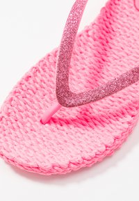 Vaaleanpunaiset flip-flopit, joissa on teksturoitu kudottu pohja. Hihna on kiiltävä ja glitteripintainen, luoden eloisan ja silmäänpistävän muotoilun.