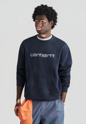 Granatowa bluza z dużym szarym logo „carhartt”, z przednią kieszenią i subtelnym śladem użytkowania. Połączona z jasnoniebieskimi spodniami i pomarańską czapką.