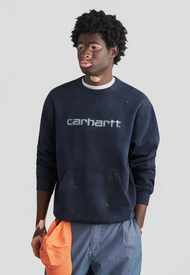Marineblauer Pullover mit großem grauem "carhartt"-Logo, mit einer Fronttasche und dezentem Gebrauchsspuren. Kombiniert mit hellblauen Hosen und einer orangenen Mütze.