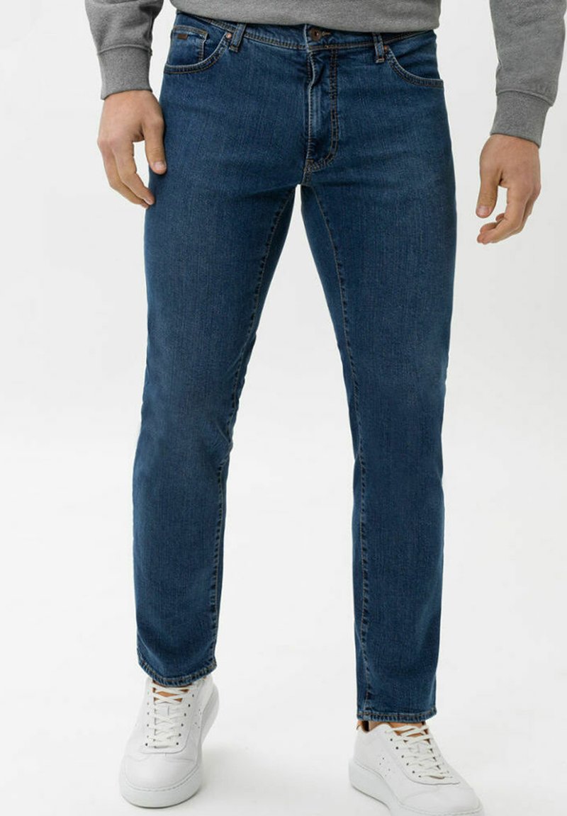 Jeans in denim blu scuro con un design a gamba dritta, cinque tasche e un delicato dettaglio di cucitura, indossati con sneakers bianche.