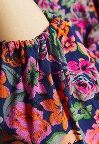 Tissu floral avec des fleurs vives en rose, orange et vert sur un fond bleu foncé. Encolure froncée avec un motif texturé.