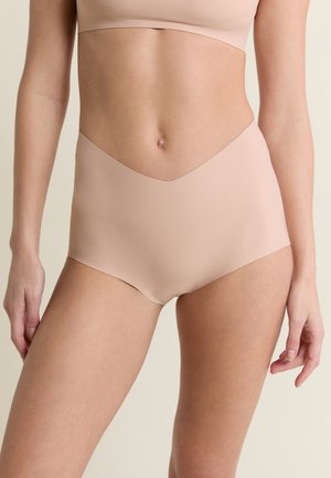 Kobieta nosząca bezszwową, cielistą bieliznę z wysokim stanem i pasujący biustonosz, stojąca na jednolitym neutralnym tle.