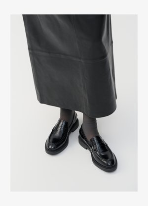 Schwarze Lederloafers mit glatter Oberfläche, verziert mit einem dekorativen Riemen, runder Zehenpartie und texturierten Sohlen. Getragen mit einem langen schwarzen Lederrock.