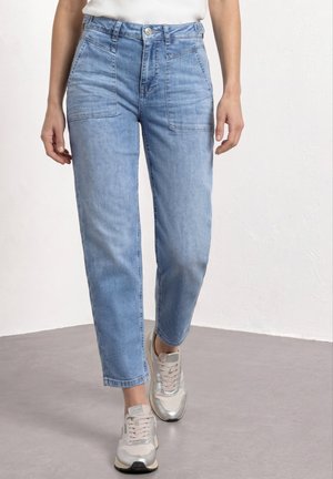 Kvinde iført lyseblå højttaljede jeans med forlommer og beige sneakers, stående på en grå gulv mod en hvid væg.