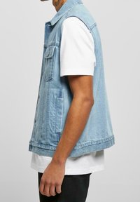 Světle modrá džínová vesta s límečkem, dvěma předními kapsami a bočními rozparky. Nenošena přes bílé tričko, ukazující volný střih.