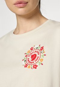 Bežinis medvilninis marškinėliai su apvaliu gėlių logotipu raudonomis kontūromis ir daugiaspalvėmis gėlėmis. Apvalus kaklas, trumpomis rankovėmis.