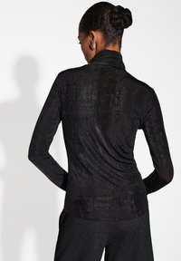 Maglietta nera a maniche lunghe con collo alto, realizzata in un tessuto testurizzato e luminoso, con un design drappeggiato sul retro. Finitura liscia e forma aderente.