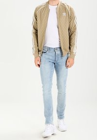 Beige bomber jacket with white polka dots, white adidas stripes on sleeves, paired with light blue jeans and white sneakers.