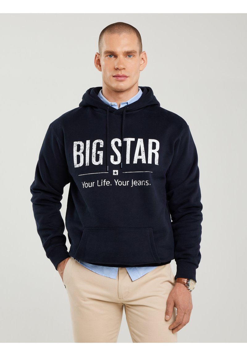 Vīrietis ar īsiem matiem, tērpies tumši zilā sporta krekliņā ar uzrakstu "BIG STAR Your Life. Your Jeans.", bēšā krāsā biksēs, zilā kreklā un ar rokassprādzi.