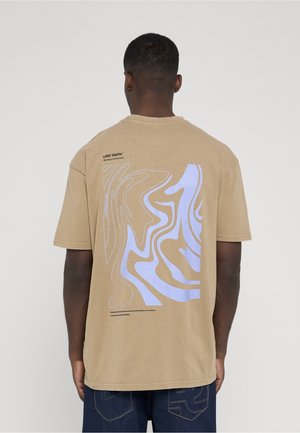 Mann trägt beige T-Shirt mit abstrakten hellblauen und weißen wellenförmigen Linien und kleinem schwarzem Text am oberen Rücken vor weißem Hintergrund.