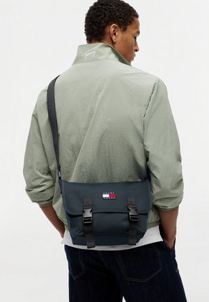 Mand i grøn jakke bærende en sort Tommy Jeans crossbody-taske med spænder, stående med hænderne i lommerne mod en ensfarvet baggrund.