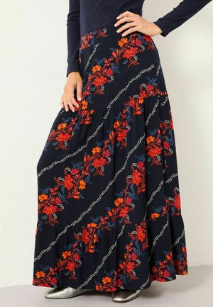 Marineblaue Maxirock mit roten und blauen Blumenmustern, gestuftem Design und wellenförmigen weißen Akzenten. Leichtes Material; silberne Schuhe ergänzen den Look.