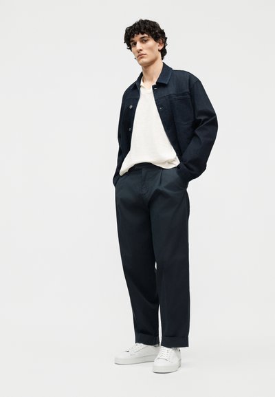 Jeune homme debout, les mains dans les poches, portant une veste en denim noire, une chemise blanche, un pantalon foncé et des baskets blanches, sur un fond uni.