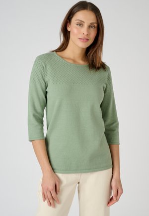 Groene gebreide trui met een gestructureerd diamanten patroon op de schouderpartij, ronde halslijn en driekwart mouwen. Gecombineerd met lichtgekleurde broek.